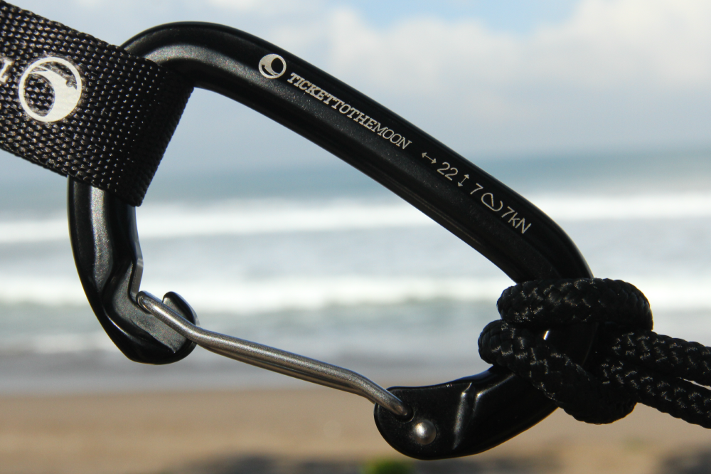 Black Carabiner 22kN (2-Pack) with 22 and 7 kN ratings engraved, clipped to a strap with the beach behind