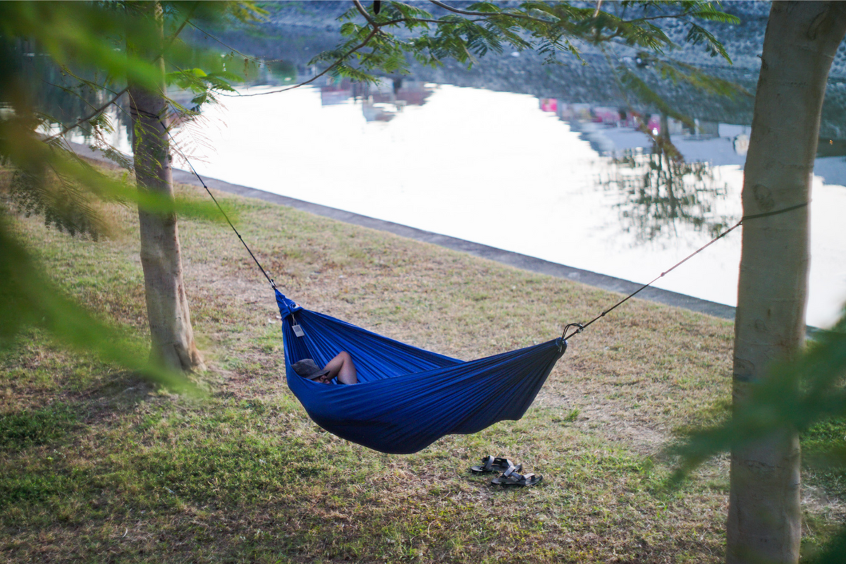 Person resting in a blue Travel Hammock Compact hung between two trees beside a river