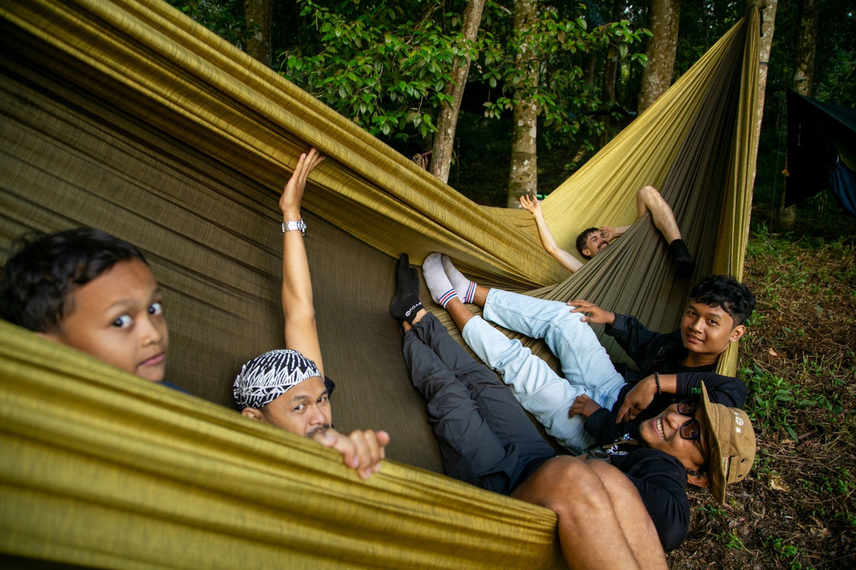 Group of friends lounging and waving inside a large olive Travel Hammock Mammock in a forest