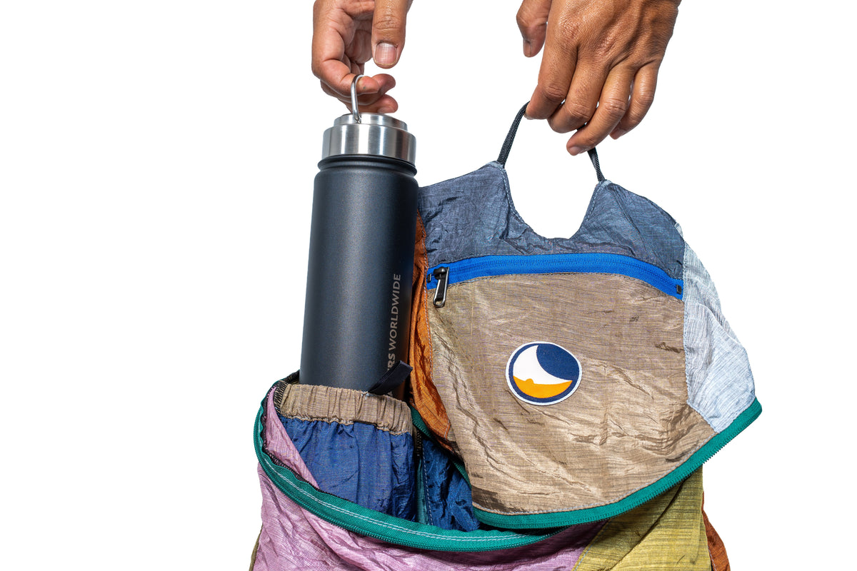 A reusable insulated bottle with a silver cap is being held into a colorful, lightweight backpack by hands against a white background. The backpack features a circular Ticket to the Moon logo.