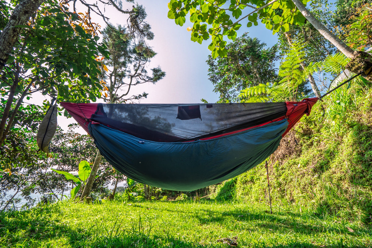 Pro Hammock with bug net and a teal underquilt, hung between trees on a grassy slope, low-angle view