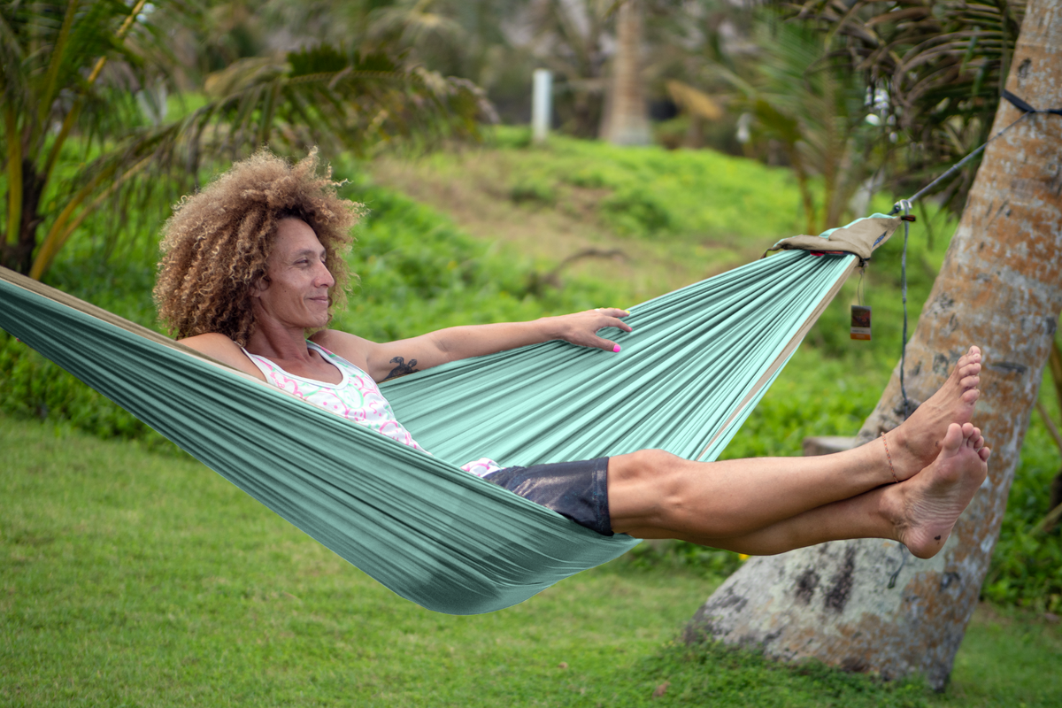 Smiling woman lounging in a mint green Travel Hammock Original tied to a palm tree, feet crossed