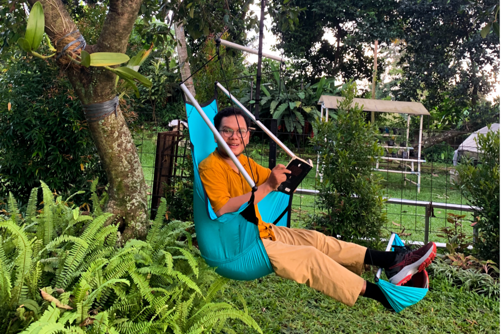 Smiling man in a turquoise Moon Chair (Custom) with footrest, hung from a tree in a leafy garden