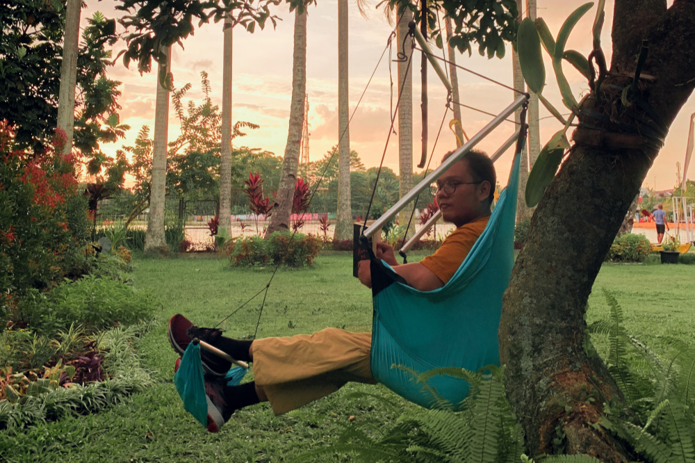 Man reading in a turquoise Moon Chair (Custom) hung from a tree in a garden at sunset