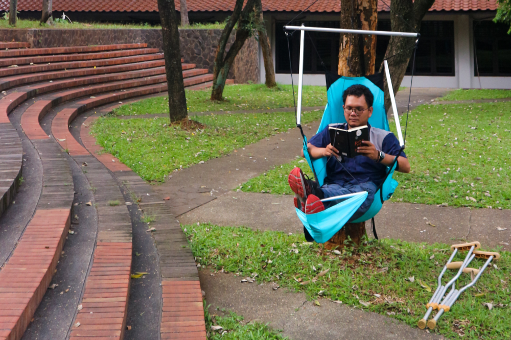Man reading a book in a turquoise Moon Chair (Custom) beside garden steps, crutches resting on the grass