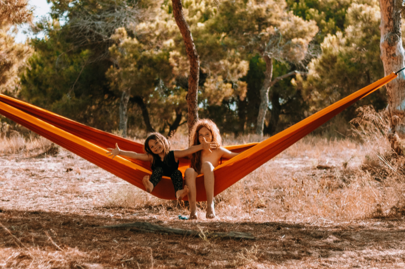 Two girls smiling in an orange Travel Hammock Honeymoon strung between pine trees in a sunny forest