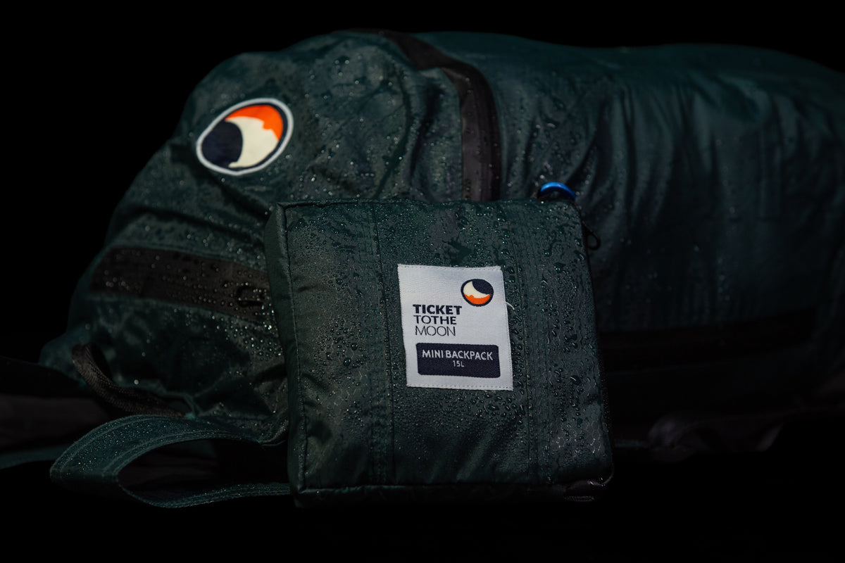 Mini Backpack 15L folded into its own pouch in front of the green backpack covered in raindrops