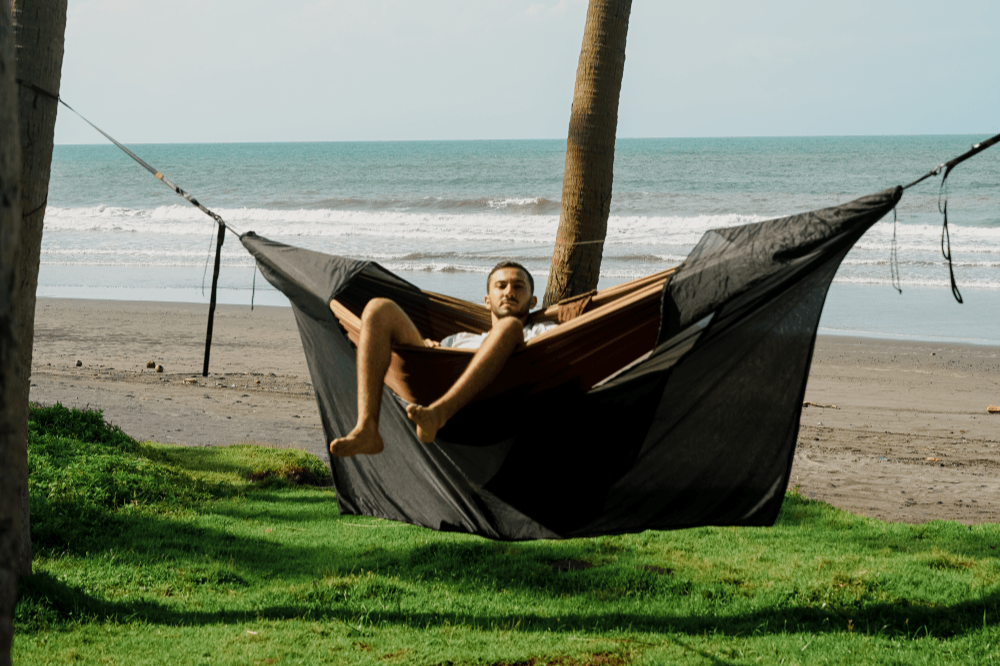 Person reclining in a hammock inside the black Universal Bug Net 360° between palm trees on a beach