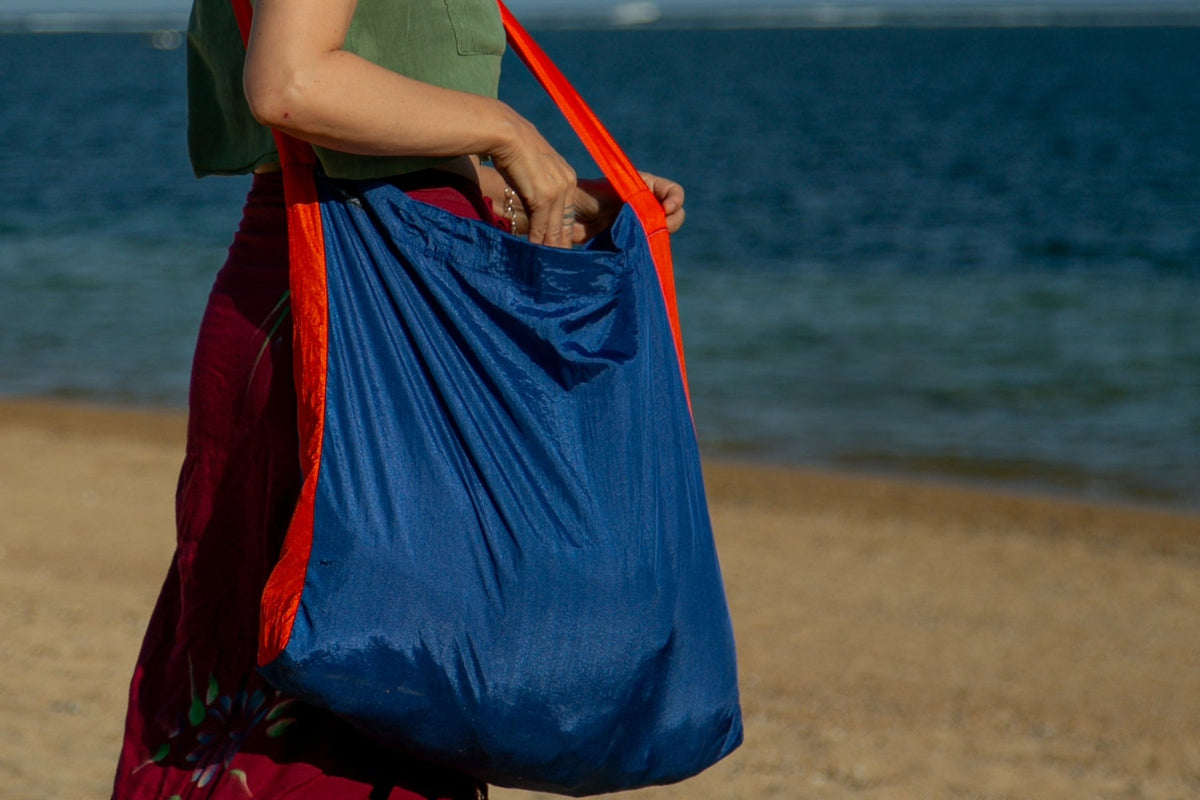 Moon Bag Large in blue with an orange strap, carried on the shoulder by a woman walking on a beach