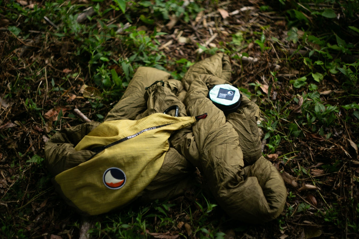 Travel Hammock Mammock in olive bundled on the forest floor beside its yellow stuff sack