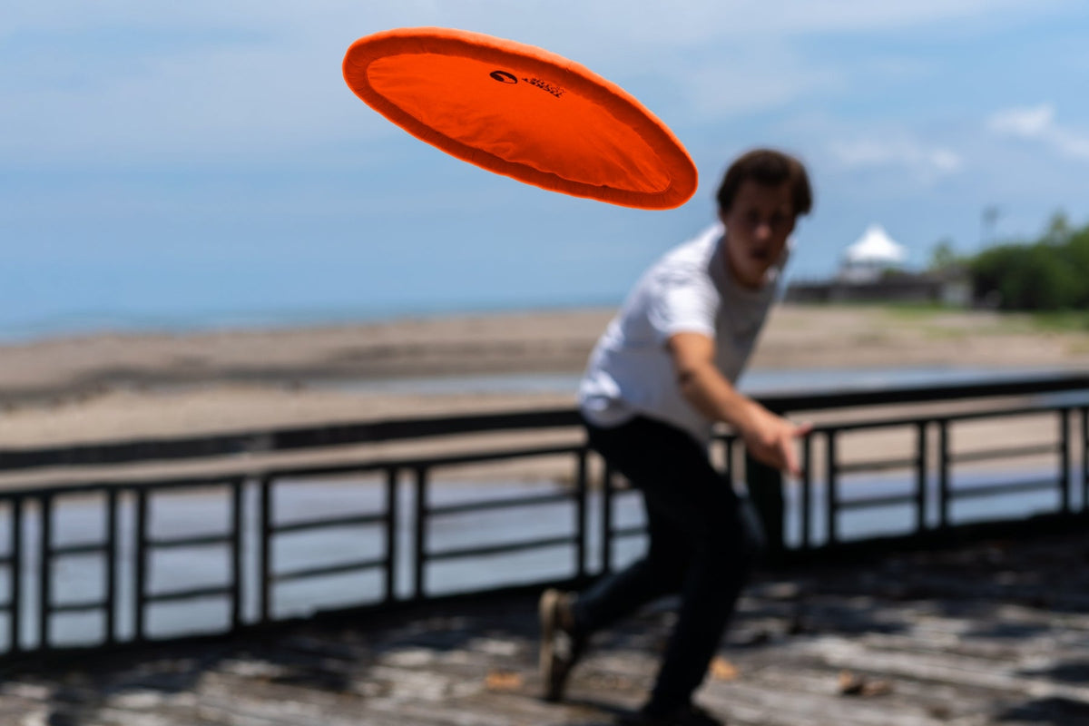 Moon Disc (Pocket) in orange flying through the air after being thrown on a seaside boardwalk