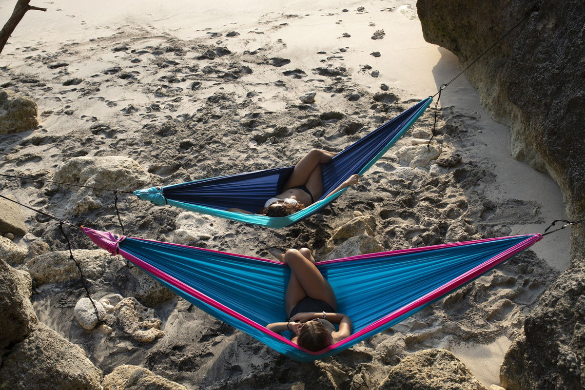 Two people relaxing in blue Travel Hammock King Size hammocks strung between rocks on a sandy beach