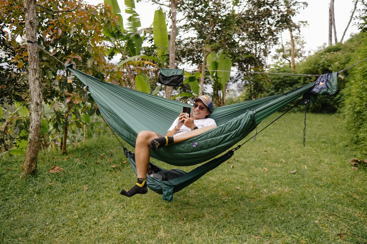 Travel Hammock Mini in green with an underquilt and a person lounging on a phone in a grassy garden