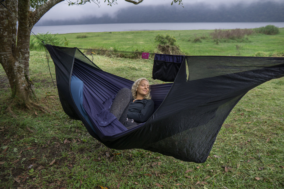 Universal Bug Net 360° in black zipped around a navy hammock with a person resting inside, on a lakeside meadow