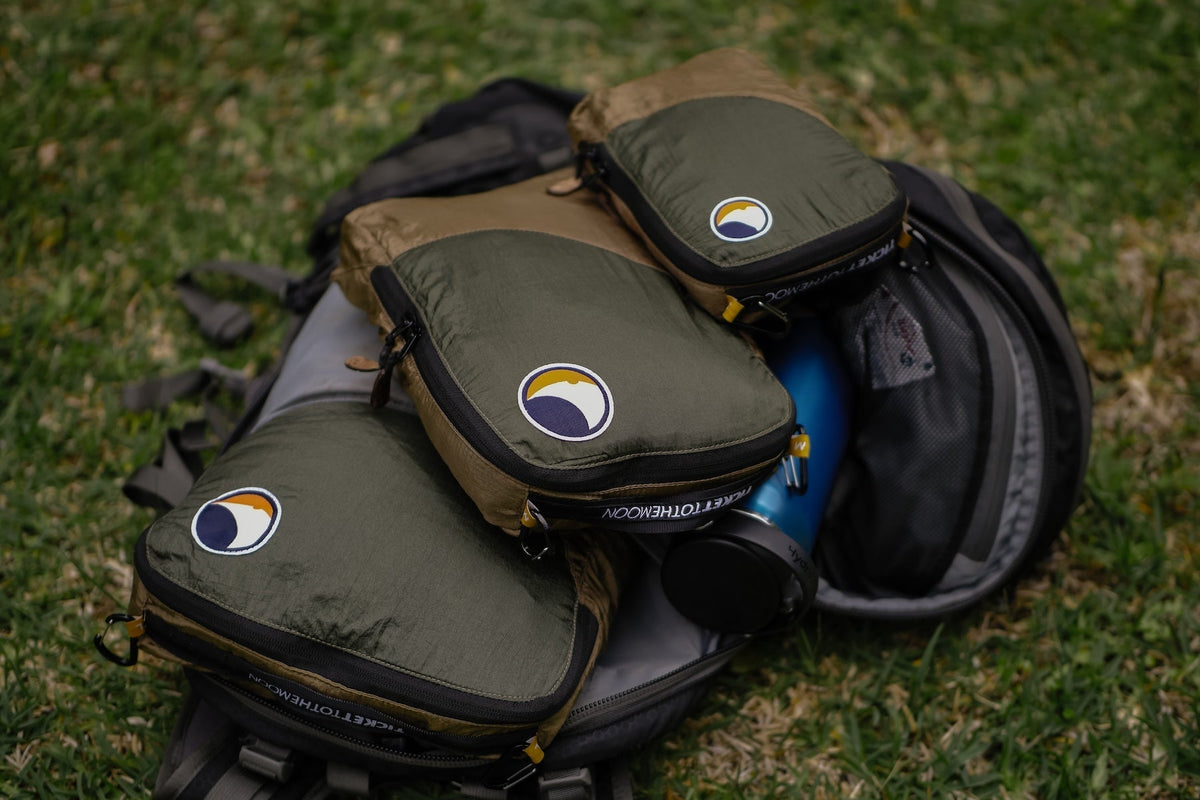 Travel Cube Set in olive green: three packing cubes stacked on an open grey backpack on the grass