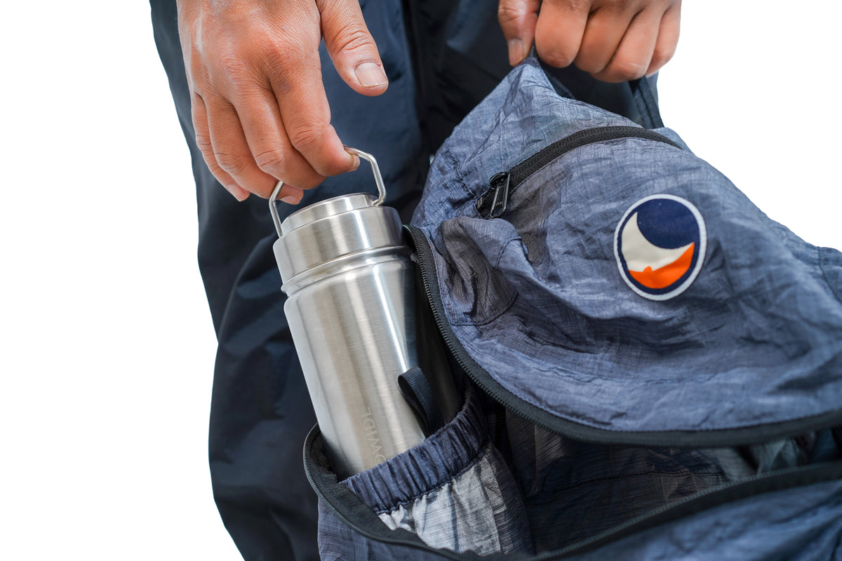 A close-up of a person's hands placing a stainless steel water bottle into the side pocket of a gray technical backpack. The backpack features a circular logo patch of Ticket to the Moon.