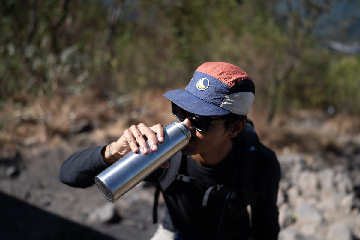 A person outdoors wearing a dark cap and sunglasses while drinking from a silver metal water bottle.