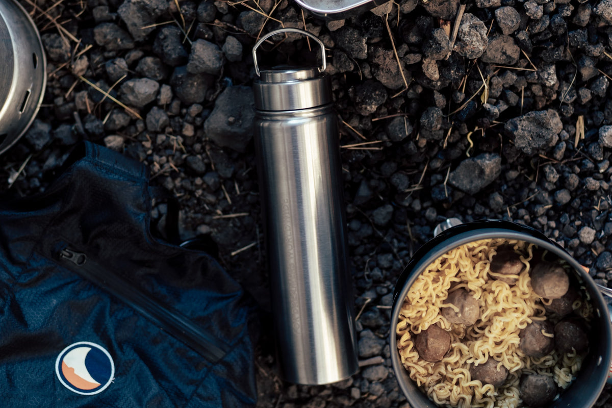Overhead view of a camping cooking setup on dark gravel ground, featuring a blue backpack, stainless steel water bottle, stove, cups, plates, and prepared food.