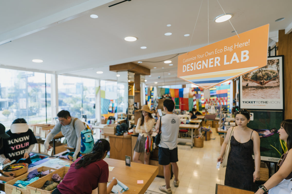 A busy retail or craft space interior with several people browsing around tables and a large orange hanging sign that reads 'Designer Lab.' The scene is indoors with bright ceiling lights, windows, and colorful merchandise displays in the background.