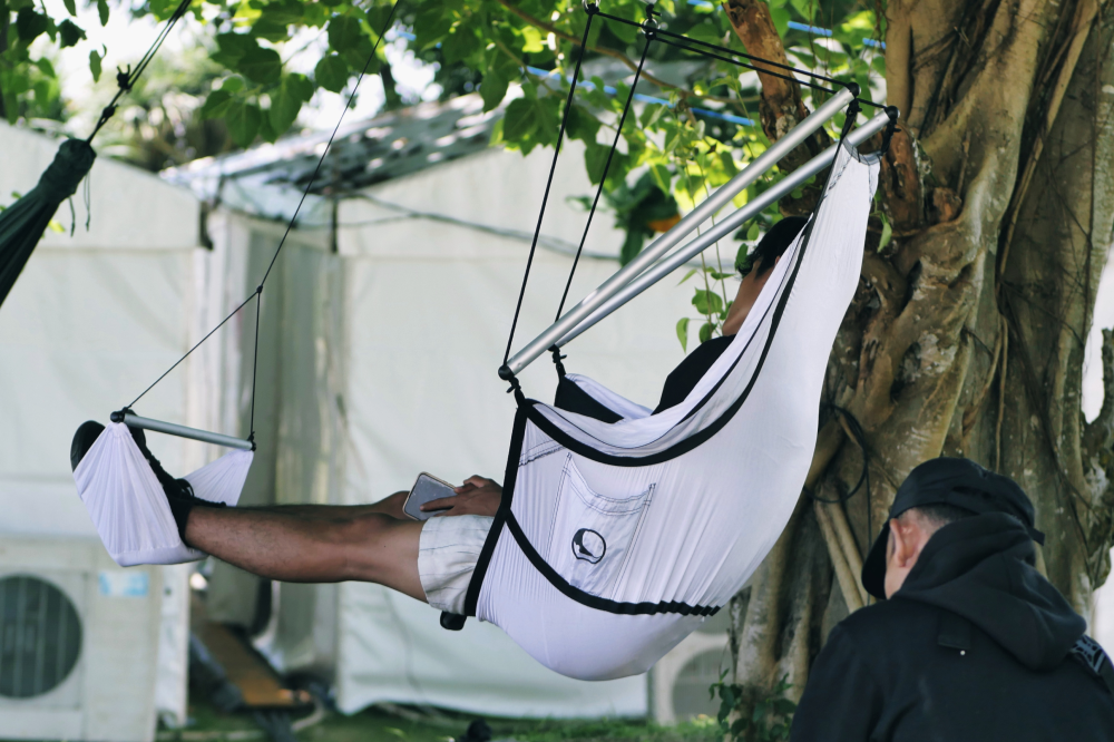 Person relaxing with a phone in a white Moon Chair (Custom) with footrest, hung from a tree at an outdoor event