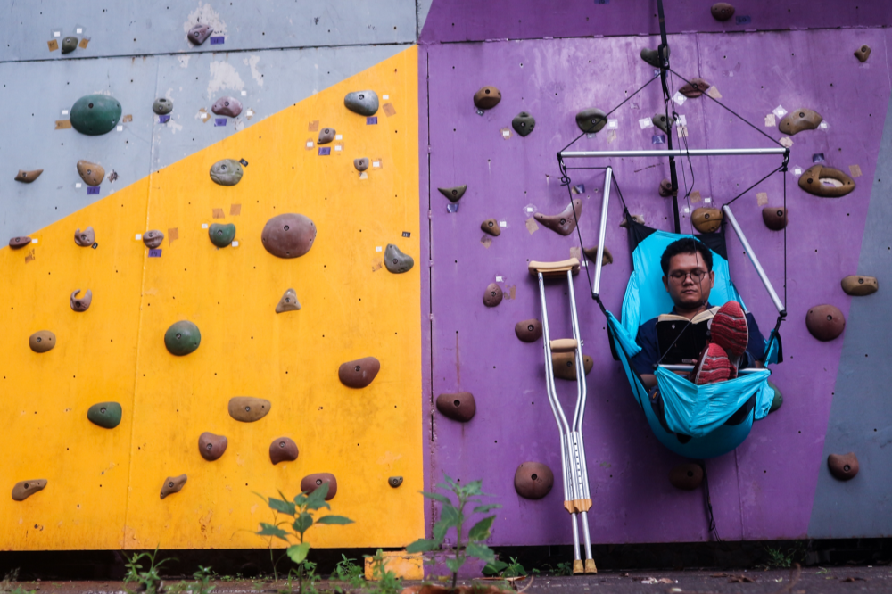 Man reading a book in a turquoise Moon Chair (Custom) hung in front of a colourful climbing wall, crutches beside him