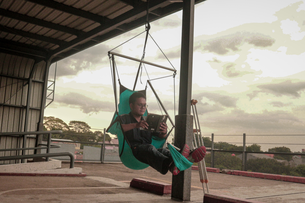 Man using a tablet in a turquoise Moon Chair (Custom) hung under a rooftop shelter, crutches nearby