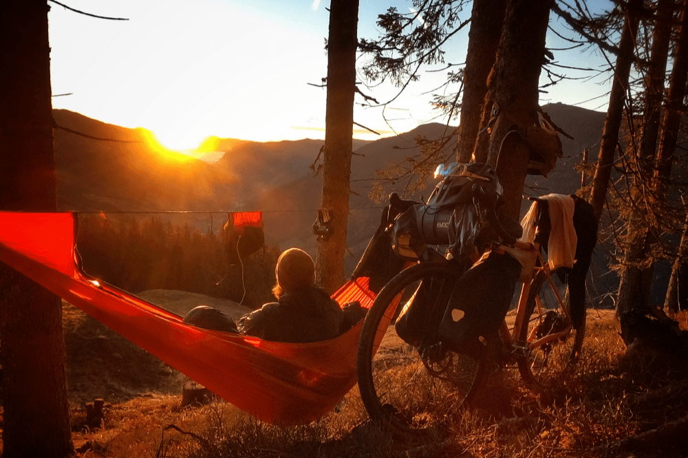 Person relaxing in an orange Pro Hammock Lightest at sunset beside a loaded touring bike in a mountain forest