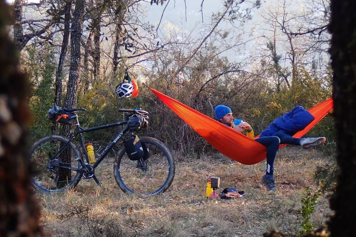 Lightest Hammock in orange hung between trees with a person reclining in it, a bicycle parked nearby