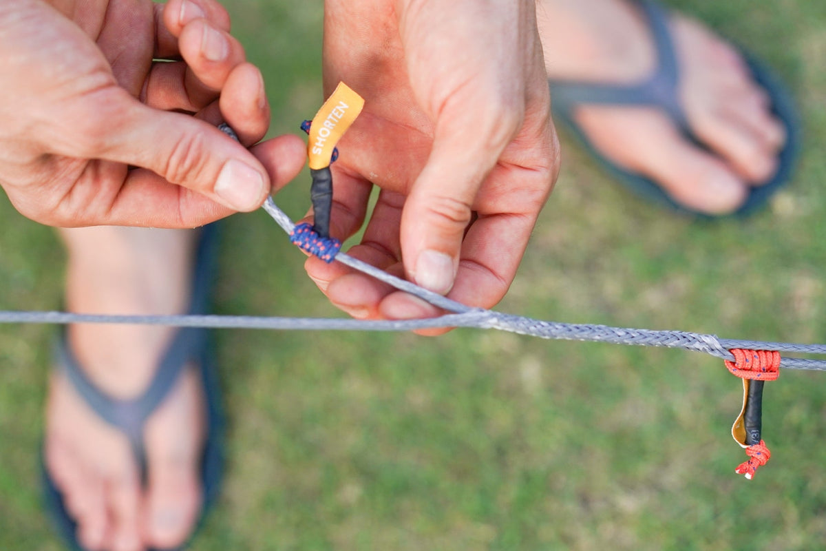 Whoopie Sling (2-Pack): hands adjusting a grey adjustable loop with an orange "Shorten" tab on a grassy background
